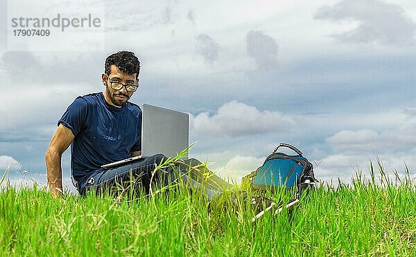 Junger Mann sitzt mit Laptop und Rucksack auf dem Feld. Mann sitzt auf der grünen Wiese und arbeitet mit seinem Laptop. Freelancer Mann Konzept arbeiten aus dem Feld