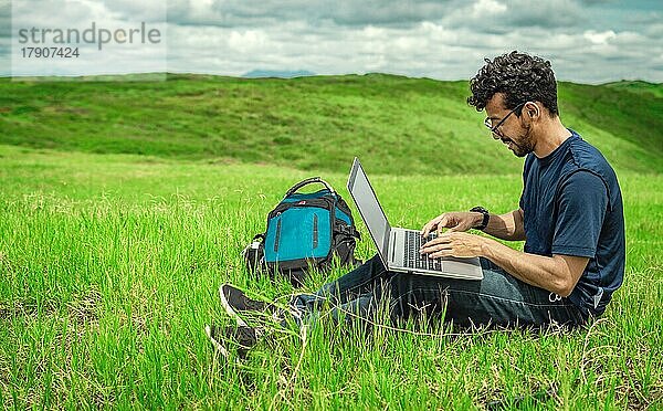 Nahaufnahme eines Mannes  der auf grünem Gras sitzt und mit seinem Laptop arbeitet. Lächelnd Freiberufler Kerl arbeiten aus dem Feld. Lächelnder Mann mit Laptop sitzt auf dem Gras auf dem Feld