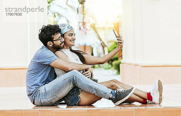 Junges Paar macht ein Selfie auf dem Boden sitzend. Junge lateinische Paar in der Liebe sitzen auf dem Boden ein Selfie im Freien zu nehmen. Konzept des glücklichen Paares  die Fotos mit dem Handy