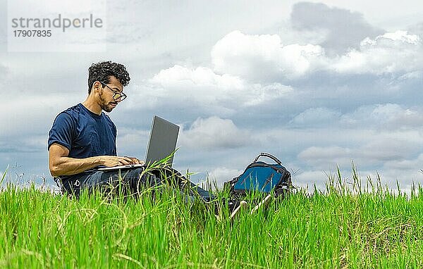 Freelancer Mann sitzt mit Laptop und Rucksack auf dem Feld. Mann sitzt auf der grünen Wiese und arbeitet mit seinem Laptop. Freelancer Mann Konzept arbeiten aus dem Feld