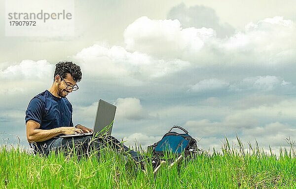 Lächelnd Freiberufler Mann sitzt mit Laptop und Rucksack im Feld. Porträt eines Mannes sitzt auf dem Gras des Feldes arbeiten mit seinem Laptop. Freelancer Mann Konzept arbeiten aus dem Feld