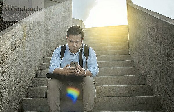 Hübscher Mann sitzt auf einer Treppe und benutzt sein Smartphone  Frontansicht eines Mannes  der auf einer Treppe sitzt und sein Handy im Freien benutzt  Nahaufnahme eines jungen Mannes