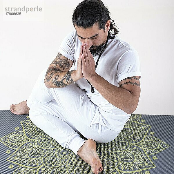 Ein weiß gekleideter Mann macht Yoga auf einer Matte vor weißem Hintergrund. Hohe Winkelansicht