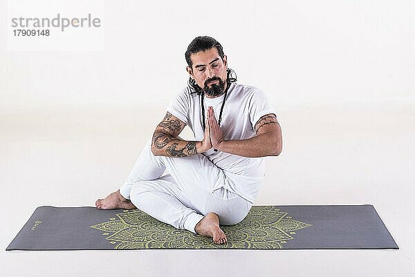 Ein weiß gekleideter Mann macht Yoga auf einer Matte vor weißem Hintergrund. Vorderansicht