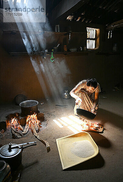 Sonnenlicht fällt in eine Küche mit Lehmwänden  Frau macht Chai  Topf auf Holzfeuer  Reisstreuer auf dem Boden  Kadana  Gujarat  Indien  Asien