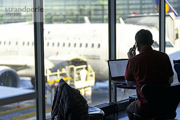 Geschäftsmann  der mit seinem Handy spricht  während er am Laptop auf dem Flughafen arbeitet  Norwegen  Skandinavien  Europa