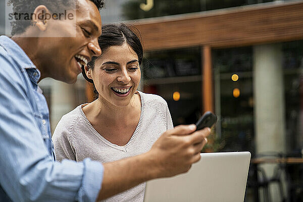 Seitenaufnahme: Afroamerikanischer Mann und lateinamerikanische Frau schauen lachend auf ein Smartphone-Display