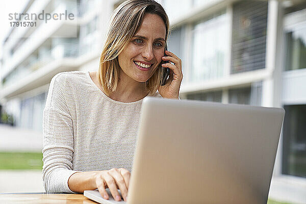Porträt einer Frau  die im Freien an einem Laptop arbeitet und mit einem Mobiltelefon spricht