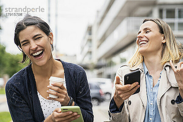 Mittlere Aufnahme von zwei Unternehmerinnen  die lachen und sich über Inhalte in den sozialen Medien freuen  während sie im Freien arbeiten