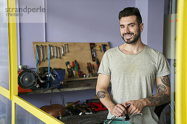 Mittelaufnahme eines lächelnden  bärtigen Mechanikers  der in seiner Werkstatt vor seinen Werkzeugen steht