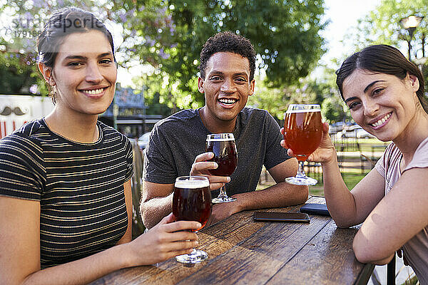 Mittlere Einstellung von drei Freunden  die in die Kamera schauen  während sie sich in einer trendigen Bar im Freien beim Biertrinken amüsieren