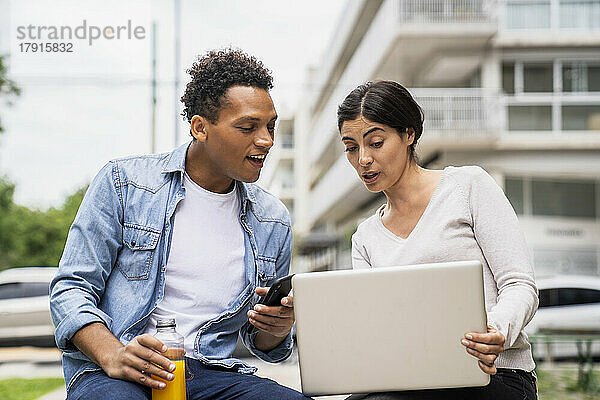 Mittelaufnahme eines afroamerikanischen Mannes und einer lateinamerikanischen Frau  die bei der Arbeit im Freien einen Laptop benutzen