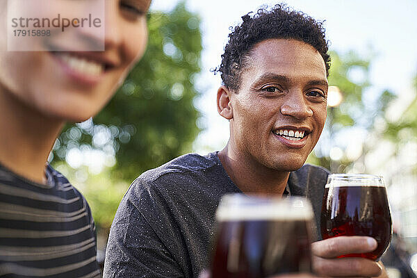 Porträt eines glücklichen jungen afroamerikanischen Mannes  der in die Kamera lächelt und ein Glas Bier hält