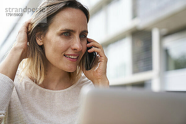 Porträt einer Frau  die vor einem Laptop sitzt und mit einem Mobiltelefon spricht