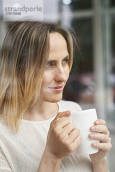 Porträt einer Frau  die eine Kaffeetasse hält und durch ein Fenster schaut