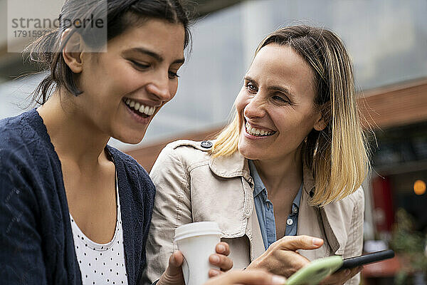 Seitenansicht von zwei Freiberuflerinnen  die im Freien arbeiten und eine gute Zeit haben  während sie ihre Smartphones überprüfen