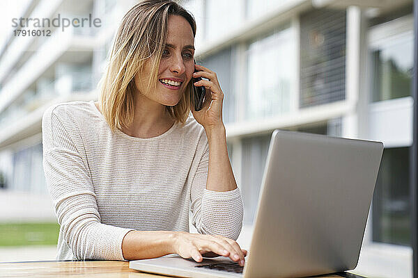 Porträt einer Frau  die im Freien an einem Laptop arbeitet und mit einem Mobiltelefon spricht