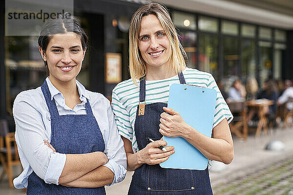 Mittlere Einstellung von zwei Restaurantbesitzerinnen  die in die Kamera schauen  während sie vor ihrem Restaurant stehen