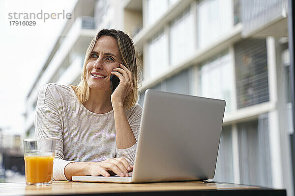 Porträt einer amüsierten Frau  die im Freien mit einem Laptop sitzt  während sie mit einem Handy telefoniert und Orangensaft trinkt
