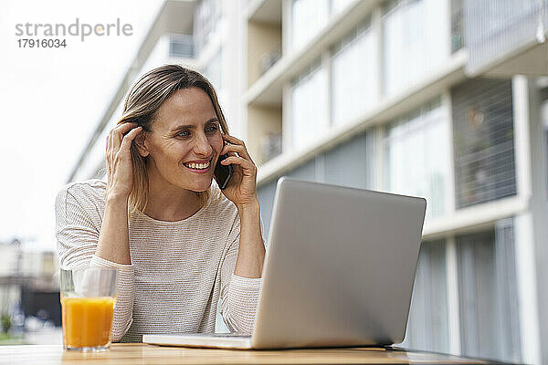Porträt einer lächelnden attraktiven Frau  die im Freien sitzt und auf einen Laptop-Bildschirm schaut  während sie mit einem Mobiltelefon spricht und Orangensaft trinkt