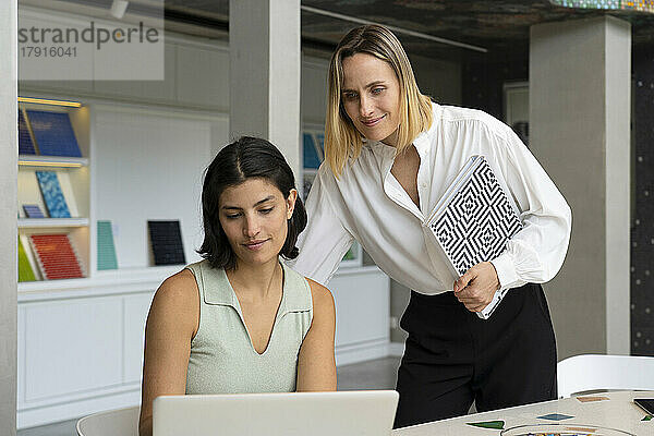 Mittelaufnahme einer Innenarchitektin und ihrer lateinamerikanischen Assistentin  die auf einen Laptop-Bildschirm schauen