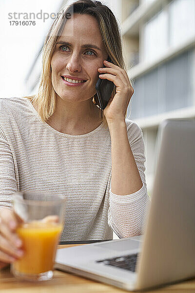 Vorderansicht einer Frau  die vor einem Laptop Orangensaft trinkt und mit einem Mobiltelefon spricht