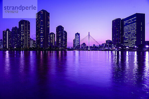 Die Stadt Tokio bei Nacht  der Sumida-Fluss  die Silhouetten der Gebäude vor dem violetten Himmel  die Spiegelungen im Wasser.