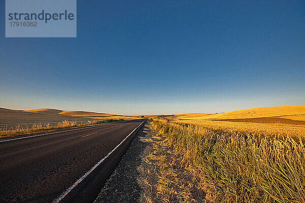 Eine Straße durch eine bäuerliche Landschaft  gerade Straße und hügeliges Ackerland  mit goldgelber reifer Weizenernte.