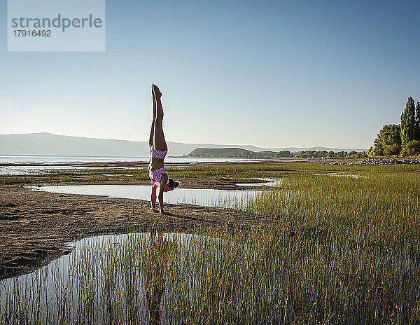 Teenager-Mädchen (14-15) im Bikini macht Handstand am See bei Sonnenaufgang