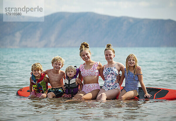 Porträt lächelnder Kinder (4-5  6-7  10-11  12-13  14-15)  die auf einem Paddelbrett am See sitzen