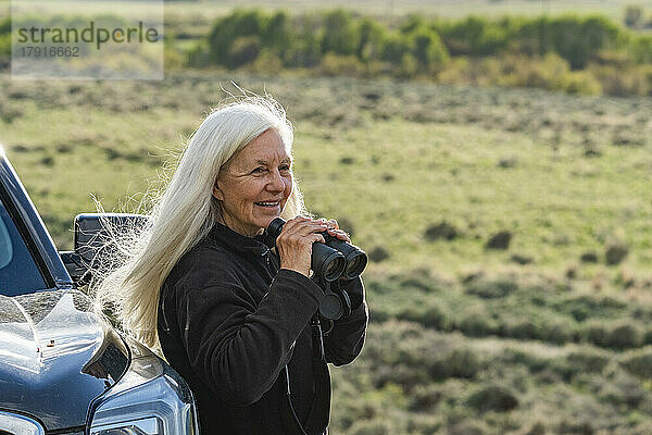 Lächelnde Frau mit Fernglas am Auto im Feld