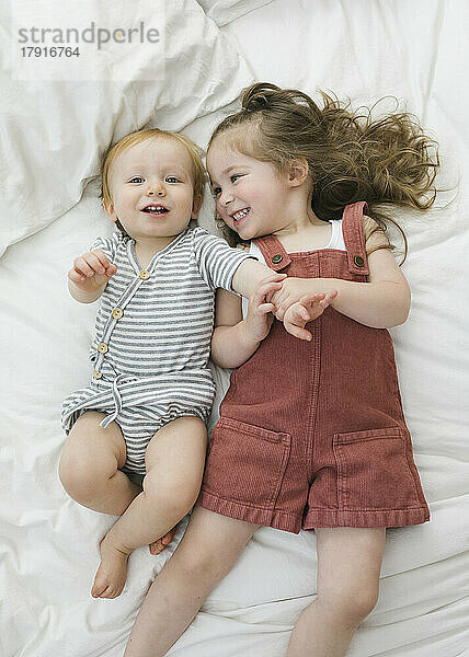 Draufsicht auf Bruder (12–17 Monate) und Schwester (2–3)  die auf dem Bett kuscheln