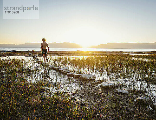 Rückansicht eines Jungen (10-11)  der bei Sonnenaufgang auf Trittsteinen im See läuft