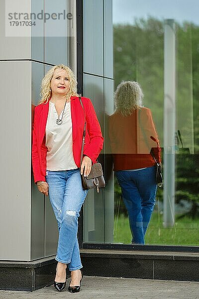 Selbstbewusste Frau in roter Jacke  Bluse und Jeans steht in der Nähe der verstärkten Glaswand des Gebäudes