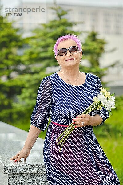 Porträt einer lächelnden reifen Frau mit kurzen rosa Haaren  die in der Nähe eines Tannenbaums auf der Straße steht