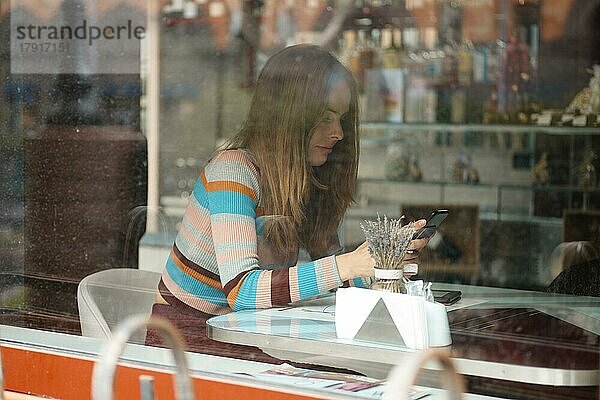 Foto durch das Fenster eines niedlichen Mädchens mit langen Haaren  das im Café sitzt und mit dem Handy telefoniert