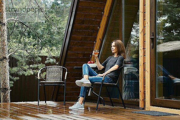 Süße Frau sitzt auf der Terrasse ihres Waldhauses und trinkt Saft