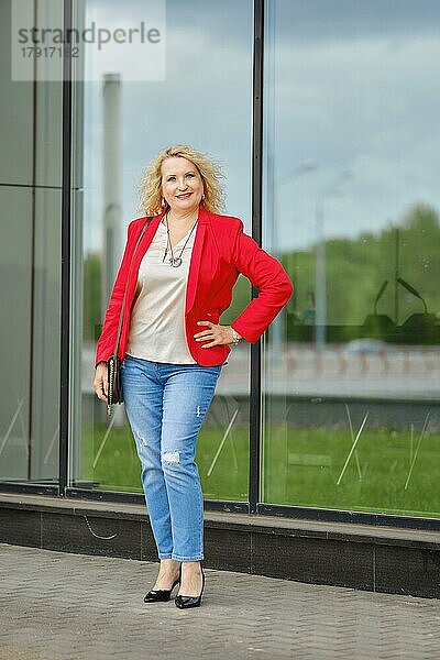 Selbstbewusste Frau in roter Jacke  Bluse und Jeans  die in der Nähe der verstärkten Glaswand des Gebäudes steht