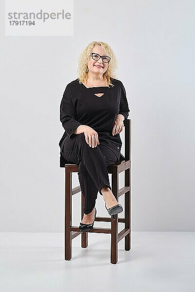 Frau mittleren Alters in schwarzem Outfit und Brille auf einem Stuhl  Studioaufnahme