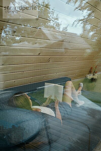 Blick durch das Fenster auf eine junge Frau auf einer Couch in einer Waldhütte  die sich mit einem Buch entspannt