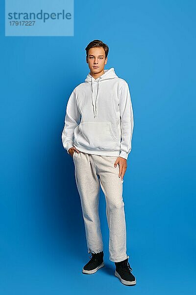 Attraktiver stilvoller Mann in weißem Sweatshirt und Jogginghose vor blauem Studiohintergrund