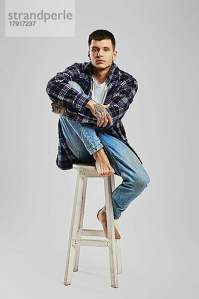 Ganzes Studioporträt eines jungen barfüßigen Mannes in Hemd und Jeans  der auf einem hohen Holzstuhl sitzt
