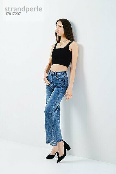 Ganzkörperporträt einer jungen Frau in schwarzem Tank-Top und Denim-Jeans  die sich an die weiße Studiowand lehnt  Seitenansicht