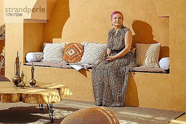 Peaceful fröhlich im Alter von Frau sitzt auf orientalischen Stil Bank mit Kissen in sonnigen Tag