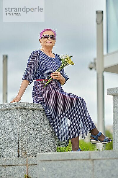 Outdoor-Portrait einer lächelnden reifen Frau mit kurzen rosa Haaren und Frühlingsblumen in den Händen