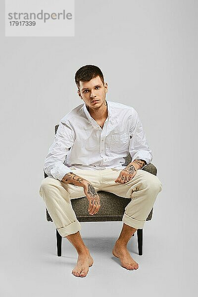 Ganzkörperporträt eines jungen barfüßigen Mannes in weißem Hemd und elfenbeinfarbenen Hosen  der in einem Sessel sitzt