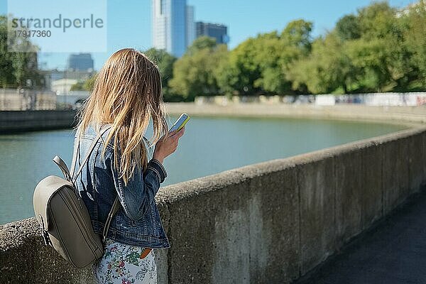 Seitenansicht eines jungen Mädchens mit Rucksack  das eine Nachricht vom Telefon liest  während es am Fluss entlang läuft