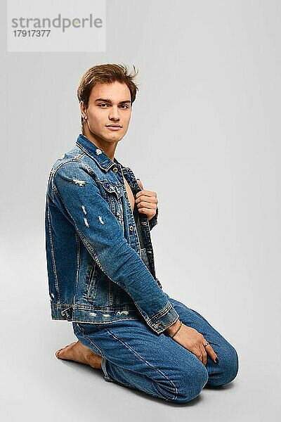 Junger Mann in Jeanskleidung sitzt auf dem Boden im Studio