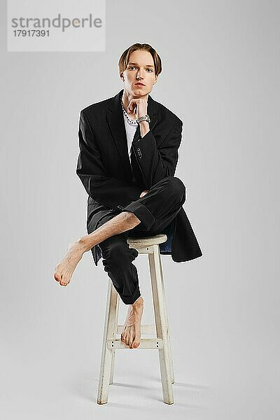 Junger barfüßiger Mann im schwarzen Anzug sitzt auf einem hohen Stuhl in einem Studio