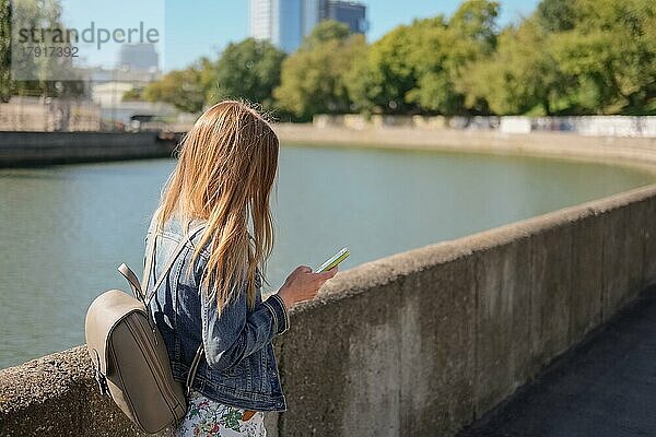 Seitenansicht eines jungen Mädchens mit Rucksack  das eine Nachricht vom Telefon liest  während es am Fluss entlang läuft
